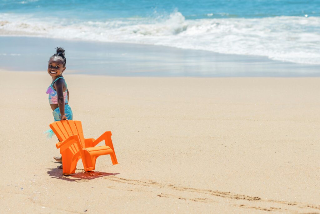 A joyful child in swimwear enjoys a sunny day at the beach, playing with an orange chair.